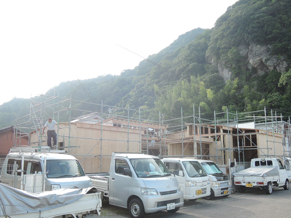 磐盾のコートハウス：緑の山を背に、足場に囲まれた上棟後の建物と工事車両