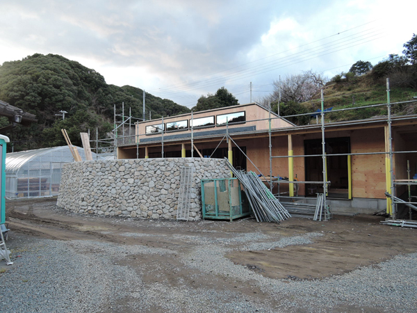 上芳養のコートハウス：石積みの塀越しに見た、構造用合板と高窓を取り付けた施工中の外観